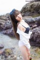 A woman in a white shirt and black bikini standing on a rocky beach.