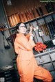 A woman in an orange jumpsuit and safety glasses in a workshop.