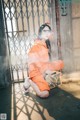 A woman in an orange jumpsuit sitting on the ground.